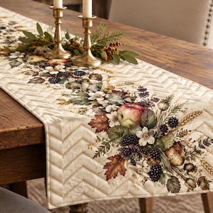 May include: A cream-colored quilted table runner featuring a central botanical print of apples, blackberries, autumn leaves, and wheat stalks. The fabric has a stitched chevron pattern throughout, creating a textured surface for dining decor.