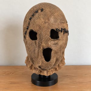Puede incluir: Una máscara de arpillera marrón con una superficie de tela tejida texturizada y detalles de costuras gruesas y oscuras alrededor de los ojos y la frente. La máscara tiene dos aberturas oculares y una gran abertura bucal ovalada sobre una base negra.