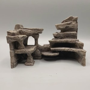 Può includere: Una struttura di formazione rocciosa testurizzata grigia e marrone con più livelli, piattaforme piatte e aperture ad arco. Il pezzo ha un aspetto stratificato e robusto con dettagli intagliati che ricordano scogliere di pietra naturale.