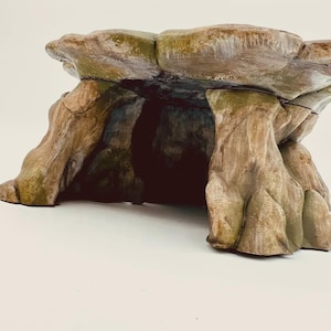Può includere: Una struttura simile a una roccia marrone e marrone chiaro con una superficie superiore ampia e piatta, sostenuta da pilastri spessi e strutturati che creano un'apertura cava a forma di grotta al centro. La superficie ha toni terrosi.