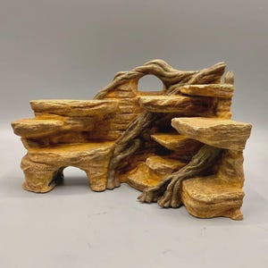 Può includere: Una formazione rocciosa testurizzata color marrone e beige con molteplici piattaforme piatte e dettagli di radici d'albero integrati. La struttura presenta un arco cavo sul lato sinistro e una piccola apertura circolare vicino alla parte superiore.
