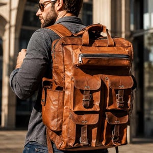 May include: A large, tan leather backpack featuring a rectangular main compartment, a top handle, and multiple exterior pockets. The front displays four smaller pockets with flap closures and metal buckles, plus a horizontal zippered pocket.