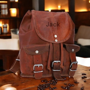 May include: A brown leather backpack featuring a top flap with the name Jack embossed in the centre. The bag includes two front pockets with silver-toned metal buckles and snap closures, along with a drawstring main compartment closure.