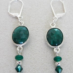 May include: A pair of silver-toned leverback earrings. Each earring features a faceted, oval-shaped, dark green gemstone set in a silver bezel. Below the oval stone, a small green bead and a faceted green crystal dangle.