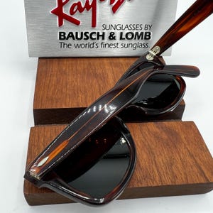 Puede incluir: Gafas de sol marrones y negras con estampado de carey. Se exhiben sobre un soporte de madera. El texto en el letrero dice: "Ray-Ban Sunglasses by Bausch & Lomb. The world's finest sunglasses."