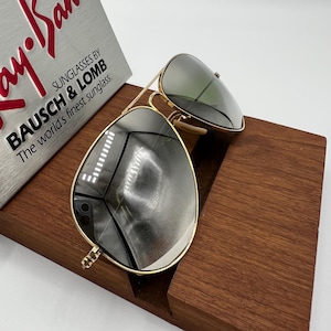 K&ouml;nnte beinhalten: Goldene Aviator-Sonnenbrille mit gr&uuml;nen Gl&auml;sern, die auf einem Holzst&auml;nder liegt. Die Sonnenbrille ist mit "Ray-Ban" und "Bausch & Lomb" beschriftet.
