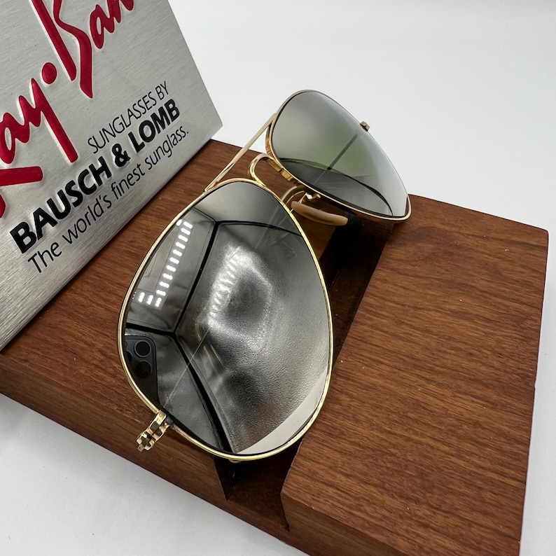 K&ouml;nnte beinhalten: Goldene Aviator-Sonnenbrille mit gr&uuml;nen Gl&auml;sern, die auf einem Holzst&auml;nder liegt. Die Sonnenbrille ist mit "Ray-Ban" und "Bausch & Lomb" beschriftet.