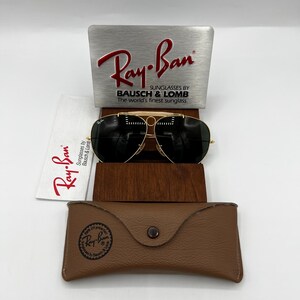 Pode incluir: &Oacute;culos de sol Ray-Ban com arma&ccedil;&atilde;o dourada e lentes escuras, apresentados com um estojo de couro castanho, um pano de limpeza e um expositor. O expositor apresenta o log&oacute;tipo Ray-Ban e o texto "Sunglasses by Bausch & Lomb".