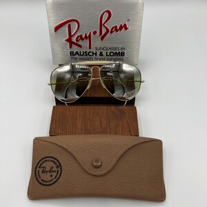 K&ouml;nnte beinhalten: Goldene Aviator-Sonnenbrille mit gr&uuml;nen Gl&auml;sern, die auf einem Holzst&auml;nder steht. Die Sonnenbrille wird mit einem braunen Lederetui mit dem Ray-Ban-Logo geliefert.