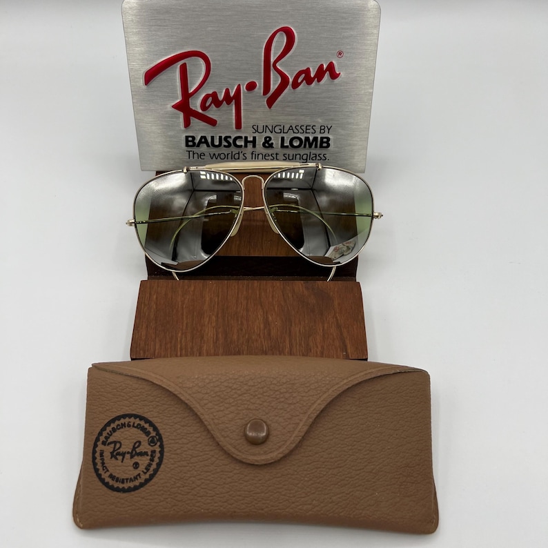 K&ouml;nnte beinhalten: Goldene Aviator-Sonnenbrille mit gr&uuml;nen Gl&auml;sern, die auf einem Holzst&auml;nder steht. Die Sonnenbrille wird mit einem braunen Lederetui mit dem Ray-Ban-Logo geliefert.