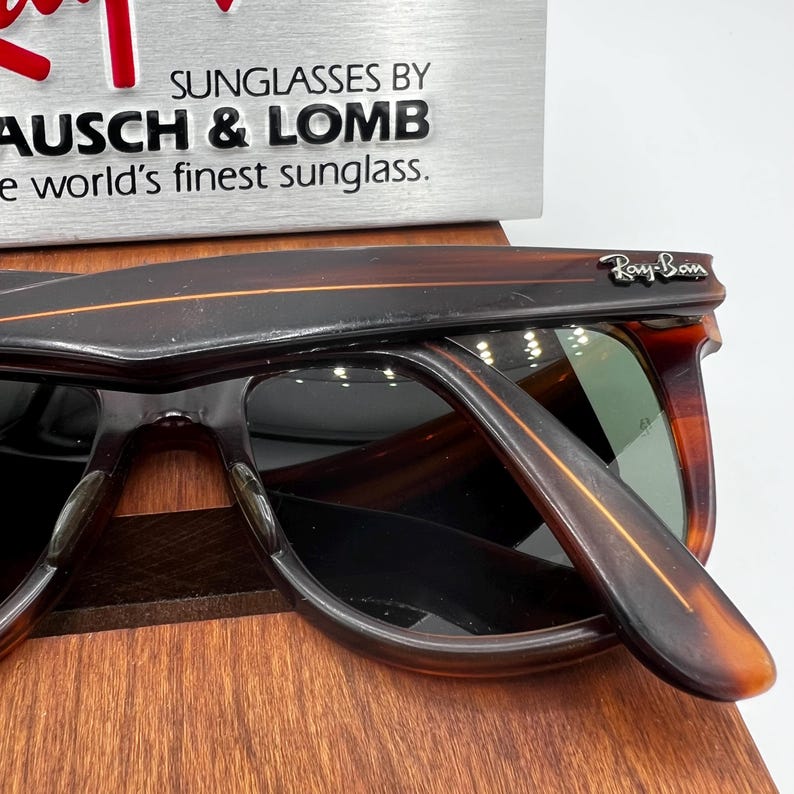 May include: Close-up of Ray-Ban sunglasses with a brown tortoiseshell frame. The sunglasses are displayed on a wooden stand. The Ray-Ban logo is visible on the temple. The text "SUNGLASSES BY BAUSCH & LOMB" is in the background.