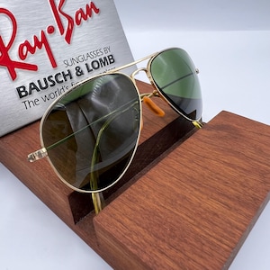 May include: Gold-framed aviator sunglasses with green lenses rest on a wooden display stand. The sunglasses are positioned in front of a silver sign with the Ray-Ban logo and the text "SUNGLASSES BY BAUSCH & LOMB".