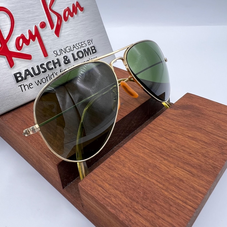 May include: Gold-framed aviator sunglasses with green lenses rest on a wooden display stand. The sunglasses are positioned in front of a silver sign with the Ray-Ban logo and the text "SUNGLASSES BY BAUSCH & LOMB".