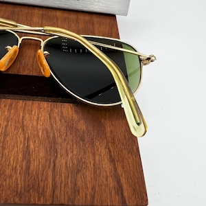 May include: Gold-rimmed sunglasses with dark lenses and a classic aviator shape. The sunglasses have a gold bridge and temples, with orange-tinted nose pads. The lenses are a dark green color. The sunglasses are resting on a wooden surface.