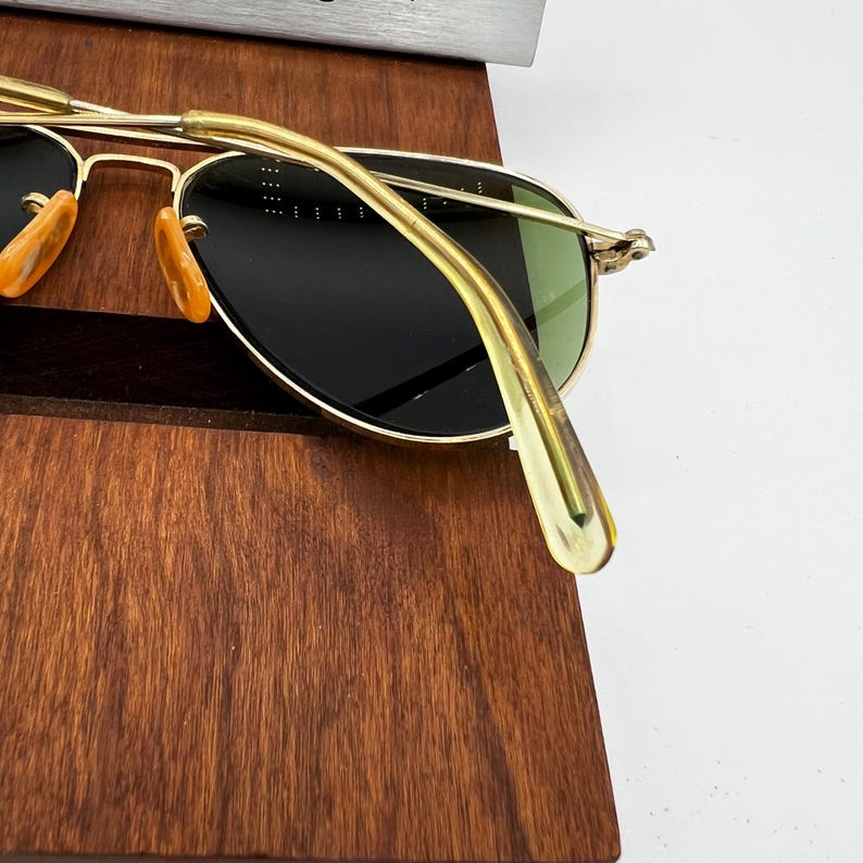May include: Gold-rimmed sunglasses with dark lenses and a classic aviator shape. The sunglasses have a gold bridge and temples, with orange-tinted nose pads. The lenses are a dark green color. The sunglasses are resting on a wooden surface.