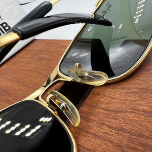 Puede incluir: Primer plano de gafas de sol con montura dorada y detalles en negro. Las lentes son de color verde oscuro y las almohadillas nasales son transparentes. La montura tiene un dise&ntilde;o elegante y moderno, y las patillas son negras con detalles dorados.
