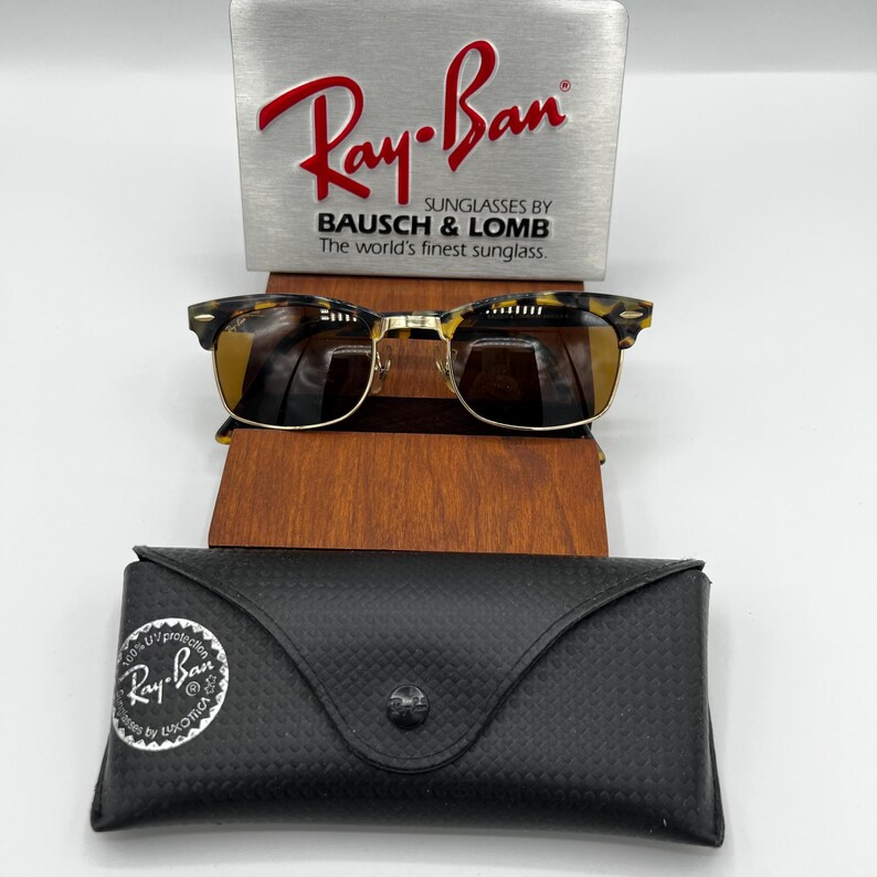 May include: Ray-Ban sunglasses with a tortoiseshell frame and gold-tone accents, resting on a wooden stand. The case is black with a textured pattern. The Ray-Ban logo is visible on the sunglasses and case.