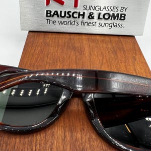 Puede incluir: Primer plano de gafas de sol Bausch & Lomb con montura marr&oacute;n oscuro con estampado de carey y lentes oscuras. El texto "SUNGLASSES BY BAUSCH & LOMB The world's finest sunglass" es visible en el fondo.