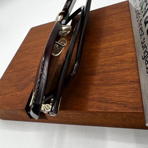 May include: Close-up of a pair of sunglasses with a dark burgundy frame and gold accents. The sunglasses are resting on a wooden display stand. The lenses are not visible. The stand is a rich, medium brown color.