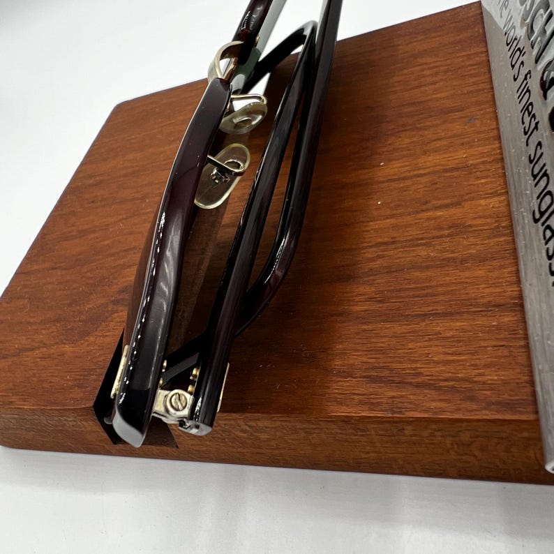 May include: Close-up of a pair of sunglasses with a dark burgundy frame and gold accents. The sunglasses are resting on a wooden display stand. The lenses are not visible. The stand is a rich, medium brown color.