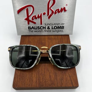Pu&ograve; includere: Occhiali da sole Ray-Ban con montatura marmorizzata verde e grigia e lenti scure. Il ponte e le cerniere dorate aggiungono un tocco di eleganza. Gli occhiali da sole sono esposti su un supporto in legno, con il logo Ray-Ban e "Sunglasses by Bausch & Lomb" sullo sfondo.