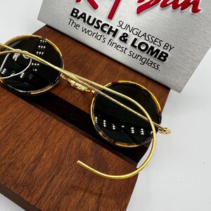 K&ouml;nnte beinhalten: Runde Sonnenbrille mit goldenem Rahmen und dunklen Gl&auml;sern auf einer Holzoberfl&auml;che. Die Sonnenbrille hat eine klassische Form und einen goldenen Metallrahmen. Der Text "BAUSCH & LOMB SUNGLASSES BY" ist sichtbar.