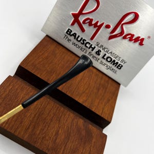 Puede incluir: Varilla de gafas de sol negra y dorada con una pieza negra para la oreja, apoyada en un expositor de madera. El logotipo de Ray-Ban es visible en un letrero plateado, con el texto "BAUSCH & LOMB Sunglasses by The world's finest sunglass."