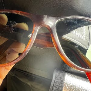 May include: Brown tortoiseshell sunglasses with dark lenses. The sunglasses have a classic frame design and are set against a dark background. The lenses appear to be slightly scratched.