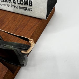 Pu&ograve; includere: Primo piano di occhiali da sole con montatura verde scuro e dettagli color oro. Il testo "BAUSCH & LOMB the world's finest sunglass" &egrave; visibile su un cartello argento e nero.