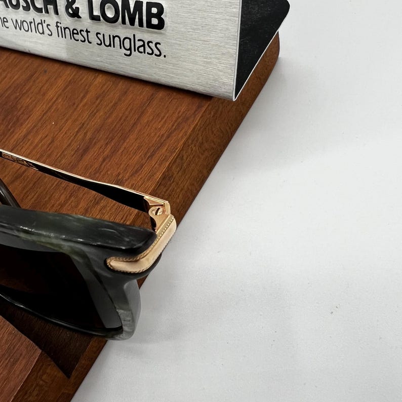 Pu&ograve; includere: Primo piano di occhiali da sole con montatura verde scuro e dettagli color oro. Il testo "BAUSCH & LOMB the world's finest sunglass" &egrave; visibile su un cartello argento e nero.