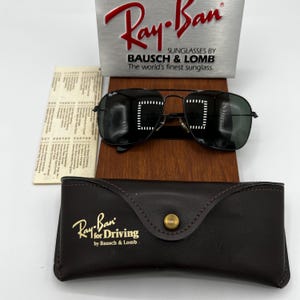 K&ouml;nnte beinhalten: Schwarze Ray-Ban Sonnenbrille mit Metallrahmen, auf einem Holzst&auml;nder. Das Bild zeigt ein braunes Lederetui und ein silbernes Schild mit dem Ray-Ban-Logo und "Sunglasses by Bausch & Lomb".