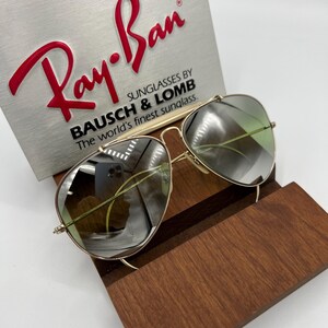 K&ouml;nnte beinhalten: Goldene Aviator-Sonnenbrille mit gr&uuml;nen Verlaufsgl&auml;sern. Die Sonnenbrille liegt auf einem Holzst&auml;nder mit einem Ray-Ban-Logo und dem Text "Sunglasses by Bausch & Lomb The world's finest sunglass."