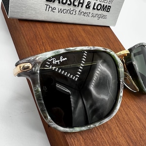 Pu&ograve; includere: Occhiali da sole Ray-Ban con montatura marmorizzata verde e grigia e lenti scure. Gli occhiali da sole hanno dettagli color oro e il logo Ray-Ban &egrave; visibile sulla lente. Il testo "The world's finest sunglass" &egrave; visibile sullo sfondo.