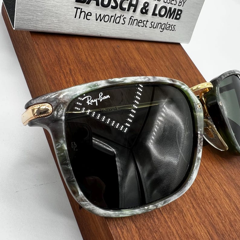 Pu&ograve; includere: Occhiali da sole Ray-Ban con montatura marmorizzata verde e grigia e lenti scure. Gli occhiali da sole hanno dettagli color oro e il logo Ray-Ban &egrave; visibile sulla lente. Il testo "The world's finest sunglass" &egrave; visibile sullo sfondo.