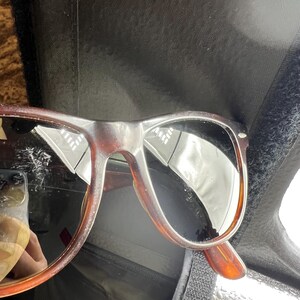 May include: Brown tortoiseshell sunglasses with dark lenses. The frames have a classic, rectangular shape with a slightly rounded edge. The sunglasses are resting on a black surface.