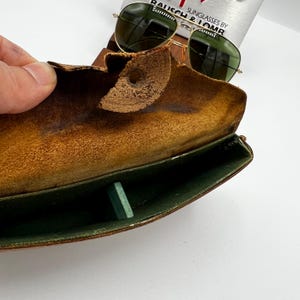 May include: Vintage gold-framed sunglasses with green lenses rest on a worn, brown leather case. The case is open, revealing a green interior and a small, rectangular green object. The text "SUNGLASSES BY BAUSCH & LOMB" is visible.