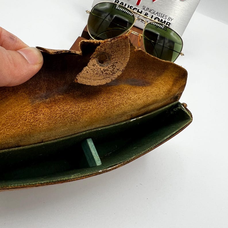 May include: Vintage gold-framed sunglasses with green lenses rest on a worn, brown leather case. The case is open, revealing a green interior and a small, rectangular green object. The text "SUNGLASSES BY BAUSCH & LOMB" is visible.