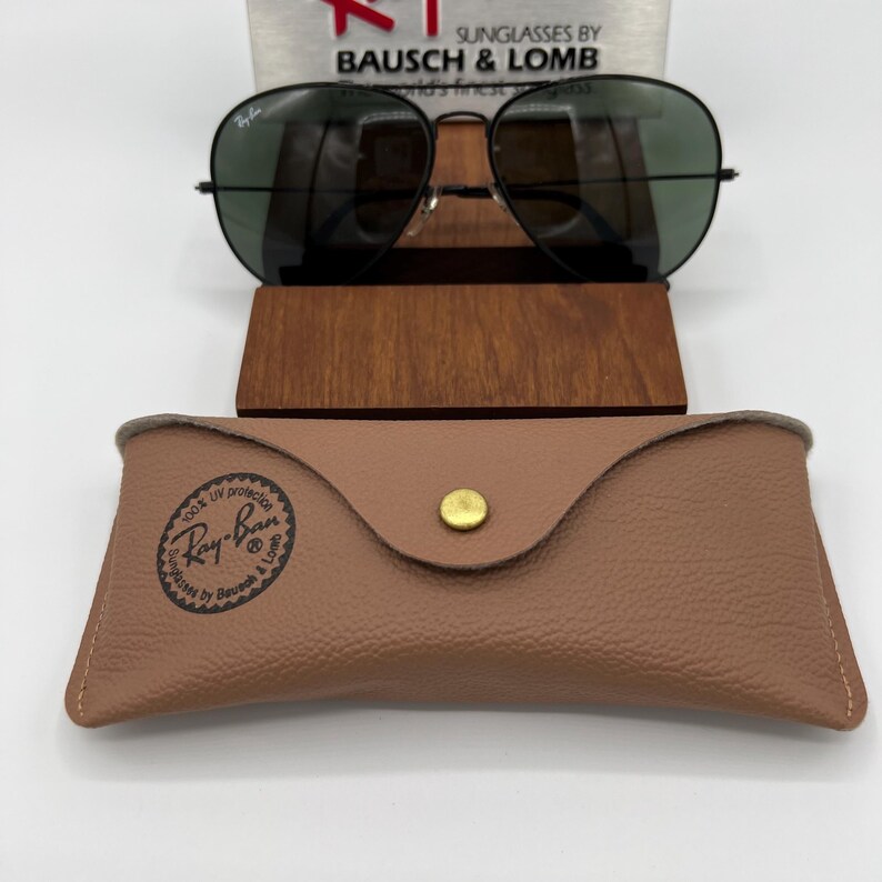 Pu&ograve; includere: Occhiali da sole aviator Ray-Ban neri con lenti verdi, in una custodia in pelle marrone con un bottone dorato. La custodia ha il logo Ray-Ban e il testo "100% UV protection Sunglasses by Bausch & Lomb".