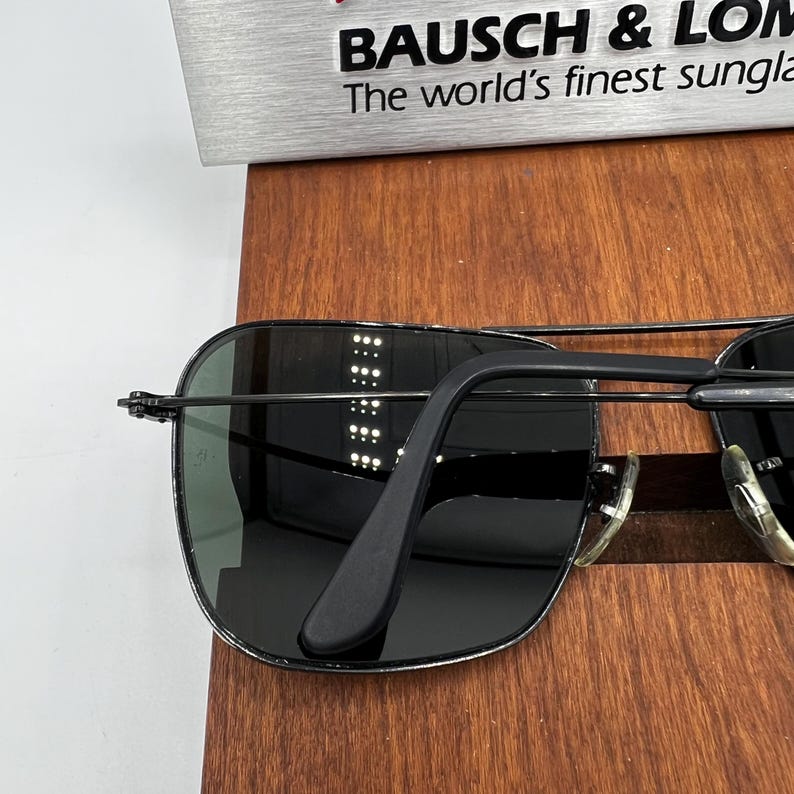 K&ouml;nnte beinhalten: Schwarze Sonnenbrille mit silbernem Rahmen und dunklen Gl&auml;sern. Die Sonnenbrille liegt auf einer Holzoberfl&auml;che. Der Text "BAUSCH & LOMB The world's finest sunglass" ist im Hintergrund zu sehen.