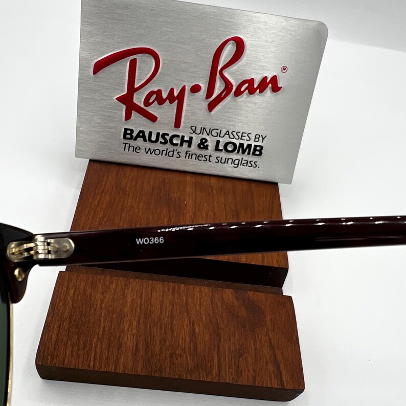 May include: Close-up of Ray-Ban sunglasses with a dark burgundy frame. The sunglasses rest on a wooden display stand. The Ray-Ban logo is in red on a silver background, with the text "SUNGLASSES BY BAUSCH & LOMB". The model number W0366 is visible.