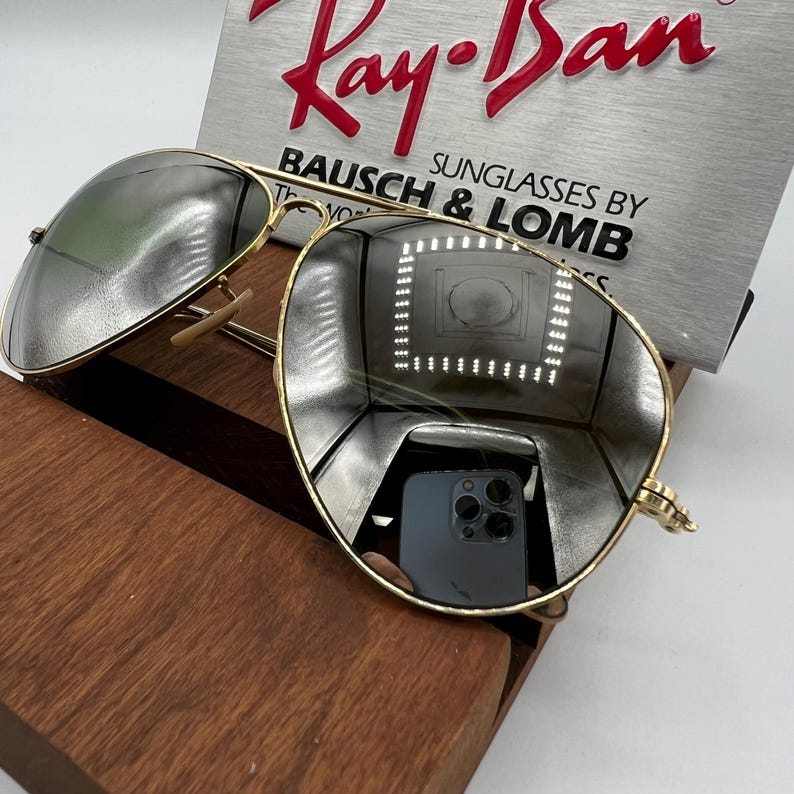 K&ouml;nnte beinhalten: Goldene Aviator-Sonnenbrille mit verspiegelten Gl&auml;sern, die ein wei&szlig;es und schwarzes Telefon reflektieren. Die Sonnenbrille liegt auf einer Holzoberfl&auml;che mit einem Ray-Ban-Etikett im Hintergrund. Sonnenbrillen von Bausch & Lomb.