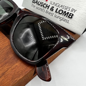 Puede incluir: Gafas de sol marrones oscuras con montura gruesa, sobre una superficie de madera. Las lentes son oscuras y la marca "BAUSCH & LOMB" es visible en un letrero plateado. Dise&ntilde;o cl&aacute;sico y atemporal.