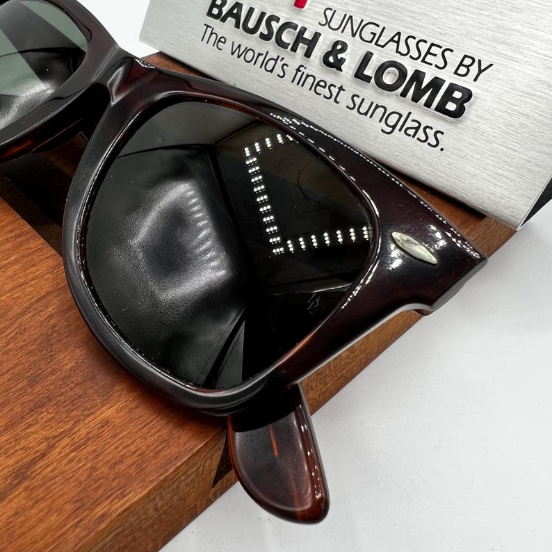 Puede incluir: Gafas de sol marrones oscuras con montura gruesa, sobre una superficie de madera. Las lentes son oscuras y la marca "BAUSCH & LOMB" es visible en un letrero plateado. Dise&ntilde;o cl&aacute;sico y atemporal.