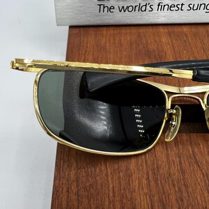 Puede incluir: Gafas de sol con montura dorada y lentes oscuras. Las gafas tienen forma rectangular y puente y varillas doradas. El texto "The world's finest sung" es visible encima de las gafas.
