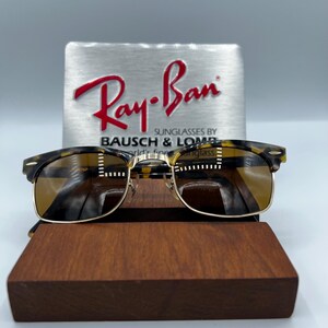 May include: Classic Ray-Ban sunglasses with a tortoiseshell frame and gold-colored accents. The lenses are a warm brown color. The sunglasses are displayed on a wooden stand, with a silver sign in the background that reads "Ray-Ban".