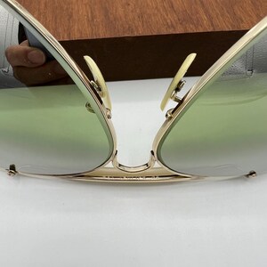 K&ouml;nnte beinhalten: Goldene Aviator-Sonnenbrille mit gr&uuml;nen Gl&auml;sern. Die Sonnenbrille ist in der Mitte gefaltet, wodurch der goldene Metallrahmen und die Scharniere sichtbar werden.