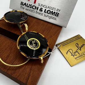 K&ouml;nnte beinhalten: Runde Sonnenbrille mit Schildpattmuster und goldfarbenen Rahmen. Die Gl&auml;ser sind dunkel, und die Sonnenbrille wird mit einem Ray-Ban-Etikett und einem Schild mit der Aufschrift "BAUSCH & LOMB, The world's finest sunglass." pr&auml;sentiert.