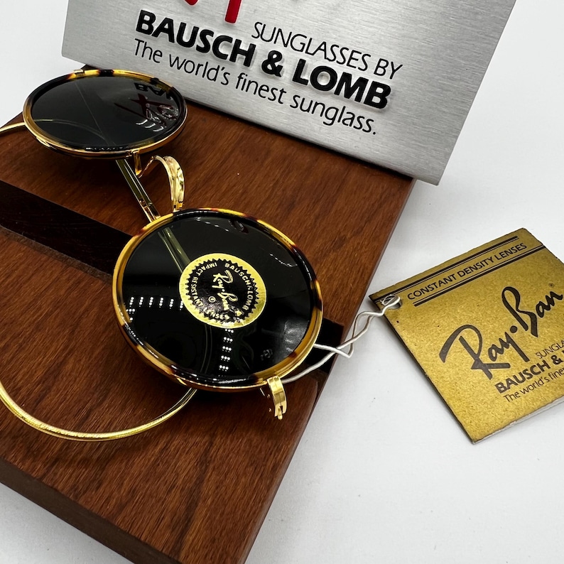K&ouml;nnte beinhalten: Runde Sonnenbrille mit Schildpattmuster und goldfarbenen Rahmen. Die Gl&auml;ser sind dunkel, und die Sonnenbrille wird mit einem Ray-Ban-Etikett und einem Schild mit der Aufschrift "BAUSCH & LOMB, The world's finest sunglass." pr&auml;sentiert.