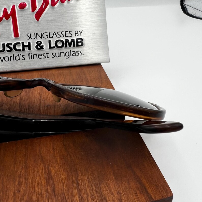 May include: Brown and black sunglasses with dark lenses. The sunglasses are resting on a wooden surface. The text "SUNGLASSES BY BUSCH & LOMB world's finest sunglass." is visible in the background.