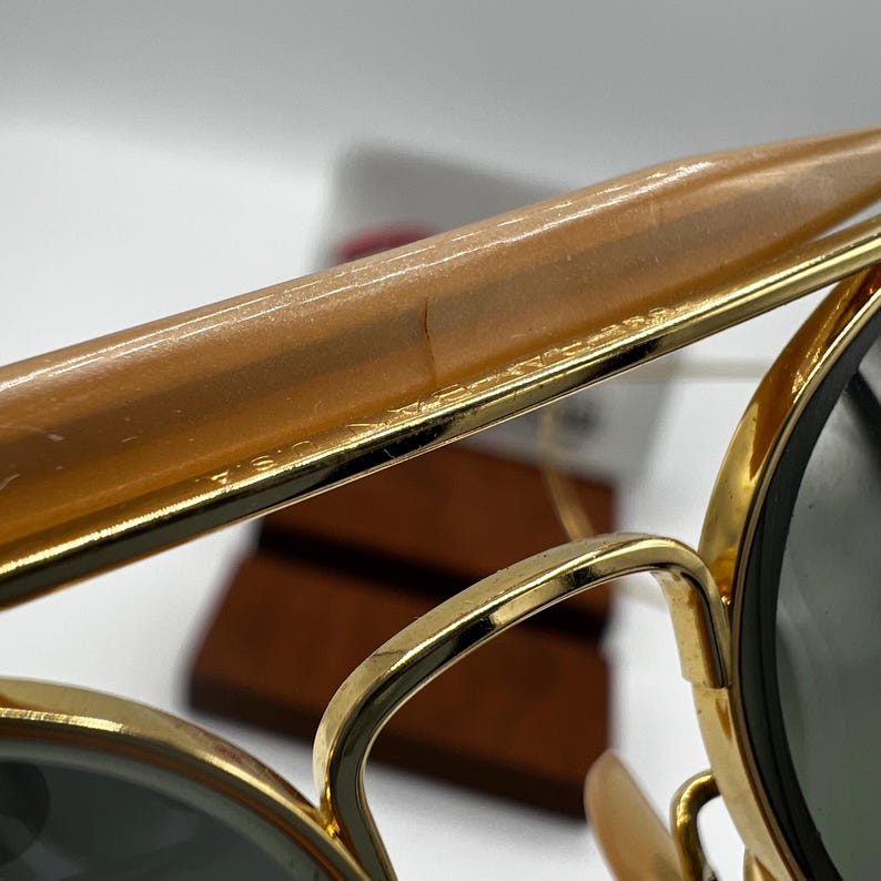 May include: Close-up of gold-rimmed sunglasses with dark lenses. The frame has a light brown top bar and the text "U.S.A." is visible on the frame. The sunglasses are set against a blurred background.